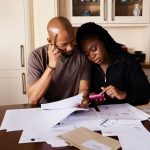 Casal sentado à mesa analisa documentos de finanças domésticas enquanto consulta um smartphone.