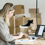 Mulher embala caixas para sua loja on-line, cercada por materiais de envio e um computador portátil.