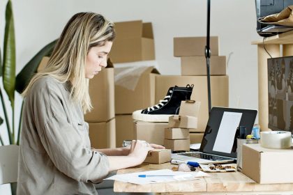 Mulher embala caixas para sua loja on-line, cercada por materiais de envio e um computador portátil.
