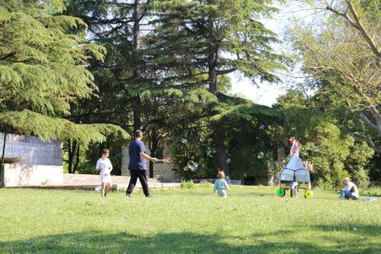 Crianças brincam com bolhas de sabão em um parque ensolarado cercado por árvores.