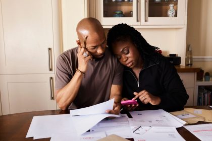 Casal sentado à mesa analisa documentos de finanças domésticas enquanto consulta um smartphone.