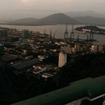 Vista aérea de uma área portuária com guindastes de carga, embarcações e casas às margens de um rio