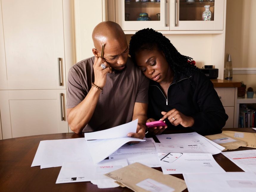 Casal sentado à mesa analisa documentos de finanças domésticas enquanto consulta um smartphone.